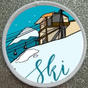May include: A white round patch with a blue and white ski scene. The scene features a ski lift, a mountain, and a ski lodge. The word "ski" is written in blue cursive lettering.