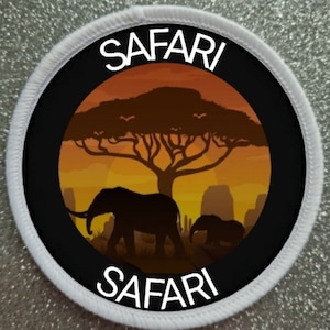 以下が含まれることがあります： サファリのシーンを描いた黒い中心部を持つ丸いパッチ。デザインには、象、木、夕日のシルエットが含まれています。「SAFARI」の文字が白で書かれており、シーンの上部と下部にアーチ状に配置され、白い縁取りが施されています。