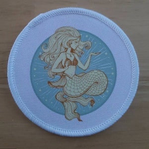 Pode incluir: Um remendo de tecido branco com um fundo azul e branco apresentando uma sereia com cabelo longo e loiro e uma cauda dourada. A sereia está segurando uma concha na mão e está cercada de estrelas.