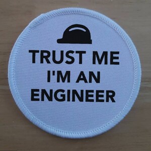 Peut inclure: Écusson rond en tissu blanc avec du texte noir qui dit "Faites-moi confiance, je suis un ingénieur" et une silhouette noire d'un casque de chantier.
