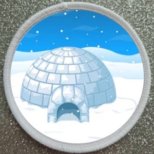 Pode incluir: Remendo redondo com um iglu de desenho animado em uma paisagem de neve. O iglu é branco e cinza, contra um céu azul com flocos de neve. O remendo tem uma borda branca costurada, para roupas ou acessórios.