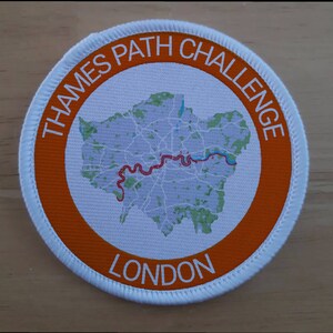 Pode incluir: Um remendo redondo branco com uma borda laranja. O remendo apresenta um mapa de Londres com uma linha vermelha que representa o caminho do Tâmisa. O texto "THAMES PATH CHALLENGE LONDON" é impresso ao redor da borda.