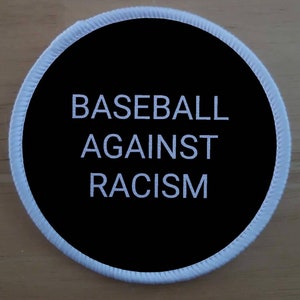Può includere: Una toppa rotonda bianca e nera con il testo "BASEBALL AGAINST RACISM" in bianco.