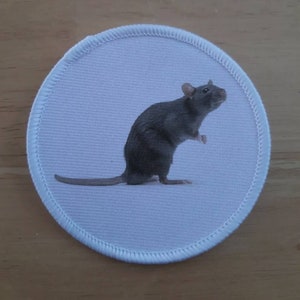 Peut inclure: Écusson rond blanc avec une illustration de rat gris. Le rat est debout sur ses pattes arrière, la queue tendue. L'écusson a une bordure blanche cousue.