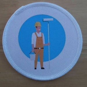 Könnte beinhalten: Ein weißer runder Aufnäher mit einem blauen Kreis in der Mitte. Der Kreis zeigt eine Cartoon-Illustration einer Person, die einen gelben Helm und eine Latzhose trägt und eine Farbrolle und einen Eimer hält.
