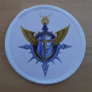 May include: White round patch with a blue and gold emblem. The emblem features a shield with a sword, wings, and a compass rose.