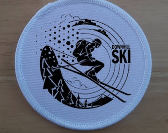 Könnte beinhalten: Ein runder weißer Patch mit einer schwarzen Silhouette eines Skifahrers, der einen Hang hinunterfährt. Der Skifahrer befindet sich in der Mitte des Patches mit einem schwarz-weißen Design eines Berges und Bäumen dahinter. Der Text "DOWNHILL SKI" befindet sich auf der rechten Seite des Patches.