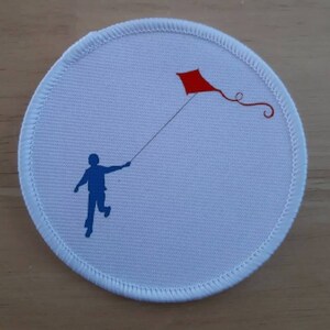 May include: A white round patch with a blue silhouette of a person flying a red kite.