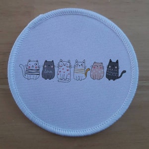 Könnte beinhalten: Ein runder weißer Stoff-Patch mit einer Reihe von sechs Comic-Katzen in verschiedenen Farben und Mustern. Die Katzen tragen Kleidung und Accessoires, darunter ein gestreiftes Hemd, eine herzförmige Halskette und ein goldenes Halsband. Der Text "love" ist auf dem Patch geschrieben.