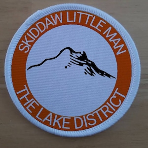 May include: White round patch with an orange border. The text "Skiddaw Little Man The Lake District" is printed in a white circle around a black silhouette of a mountain.