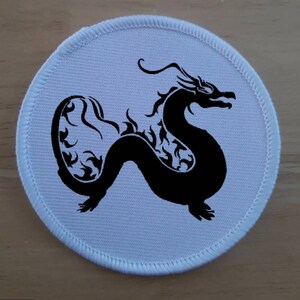 May include: A white round patch with a black silhouette of a dragon with intricate details. The dragon is facing right and has a long tail.