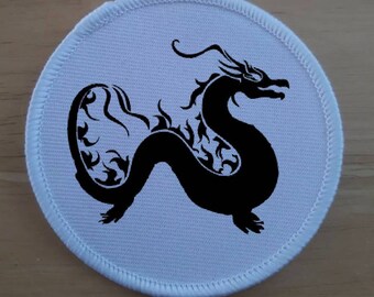 Può includere: Un cerotto rotondo bianco con una silhouette nera di un drago con dettagli intricati. Il drago è rivolto a destra e ha una lunga coda.