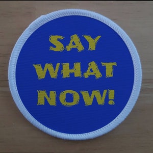 Puede incluir: Un parche redondo azul con costuras blancas alrededor del borde. El texto "SAY WHAT NOW!" está impreso en amarillo.