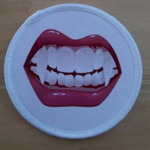 May include: A round, white patch with a stitched border. The patch features a graphic of a mouth with bright red lips and a wide, toothy grin. The teeth are white, and the lips are glossy.