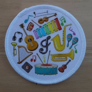 May include: A white round patch featuring a colourful design of various musical instruments, including a guitar, trumpet, drums, xylophone, and maracas. The patch has a white border with a stitched edge.