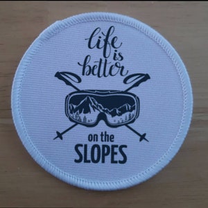 Puede incluir: Parche redondo blanco con texto negro y un gráfico de gafas de esquí y bastones de esquí cruzados. El texto dice "life is better on the SLOPES".