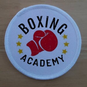 Puede incluir: Parche redondo blanco con texto negro que dice "Boxing Academy" y un gráfico de guante de boxeo rojo. Hay cuatro estrellas amarillas alrededor del gráfico.