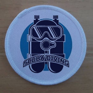 Può includere: Toppa rotonda bianca con un logo di immersioni subacquee blu e bianco. Il logo presenta una bombola da sub con una maschera e il testo "SCUBA DIVING".