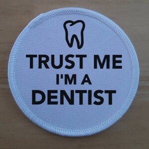 May include: White round fabric patch with black text and a tooth graphic. The text reads "Trust me I'm a Dentist".