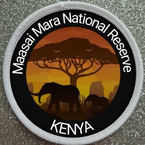 May include: A white circular patch with black text and a black circle in the center. The text reads "Maasai Mara National Reserve" and "KENYA". The black circle features a silhouette of an acacia tree with two elephants in the foreground against a sunset background.