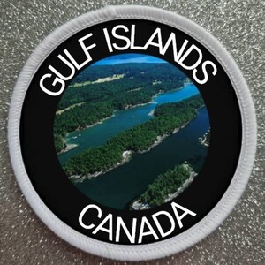 May include: A white and black round patch with the text "GULF ISLANDS CANADA" in white lettering. The patch features a photo of a body of water surrounded by green trees.
