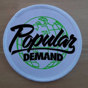 Puede incluir: Parche redondo blanco con un globo terráqueo verde y el texto "Popular Demand" en letras cursivas negras.