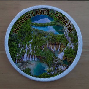 May include: A round white patch with a colourful image of Plitvice Lakes in Croatia. The text "PLITVICE LAKES - CROATIA" is written in a gold font around the edge of the patch.