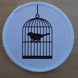 May include: A white round fabric patch with a black silhouette of a bird inside a cage.
