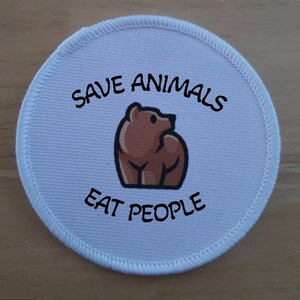May include: A round, white patch with a stitched border. The patch features a brown bear illustration and the text "SAVE ANIMALS" above and "EAT PEOPLE" below in black, bold font. The patch is likely an iron-on or sew-on.