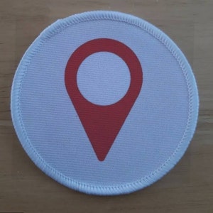 May include: A white round fabric patch with a red location pin icon in the center.