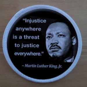 May include: A black and white round patch with a white border. The patch features a quote by Martin Luther King Jr. that reads "Injustice anywhere is a threat to justice everywhere."