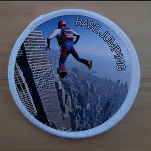 May include: A white round patch with a blue background and a picture of a person base jumping from a tall building. The text "BASE JUMPING" is written in black around the edge of the patch.