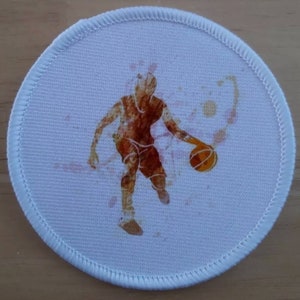 May include: A white round patch featuring a brown silhouette of a basketball player dribbling a basketball. The background is a watercolour-style design with orange and pink splatters.