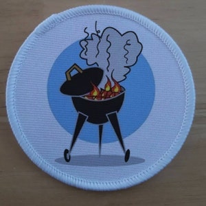 Può includere: Un cerotto rotondo bianco con un cerchio blu al centro. Il cerchio presenta un'illustrazione di cartone animato di una griglia nera con fiamme e fumo che escono da essa.