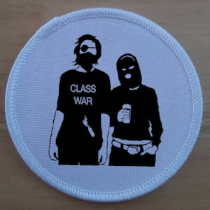 Könnte beinhalten: Schwarz-weiß-Patch mit zwei Figuren in schwarzer Kleidung. Die Figur links trägt eine schwarze Maske und den Text "CLASS WAR" auf ihrem Shirt. Die Figur rechts trägt eine schwarze Sturmhaube und hält eine Dose.
