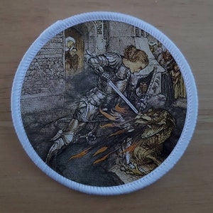 May include: A round embroidered patch featuring a knight in armor slaying a dragon with a sword. The knight is wearing a helmet and chainmail armor. The dragon is green and has flames coming from its mouth. The background is a medieval scene with a stone building and people watching.
