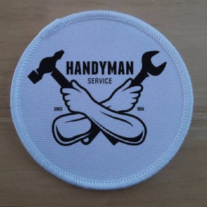 Pode incluir: Um remendo redondo branco com costuras pretas ao redor da borda. O remendo tem uma ilustração em preto e branco de ferramentas cruzadas, um martelo e uma chave inglesa, com o texto "HANDYMAN SERVICE" e os anos "SINCE 1984" impressos nele.