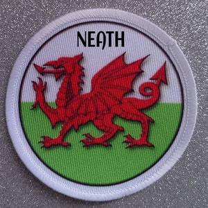 Peut inclure: Un écusson circulaire blanc avec un dragon rouge sur un fond vert et blanc. Le texte "NEATH" est imprimé en noir en haut de l'écusson.