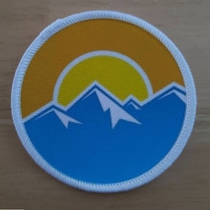 May include: A round, white-bordered patch featuring a blue mountain range silhouette with a yellow sun rising above the peaks.