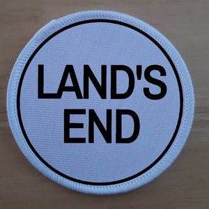 May include: White round fabric patch with black stitching around the edge. The patch has black text that reads "LAND'S END" in all capital letters.