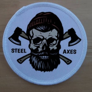 Puede incluir: Parche redondo blanco con una calavera en blanco y negro con barba y un gorro rojo y blanco. Dos hachas cruzadas están detrás de la calavera. El texto "STEEL AXES" está impreso en el parche.