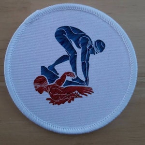 May include: A white round patch with a blue and red silhouette of two swimmers. The swimmer on top is in a diving position, while the swimmer on the bottom is in a swimming position.