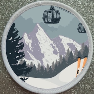 Op de afbeelding: Een ronde witte patch met een winterscène. De patch toont een besneeuwde bergketen met een bos op de voorgrond. Twee kabelbanen zijn zichtbaar in de lucht, en een paar ski's liggen in de sneeuw.