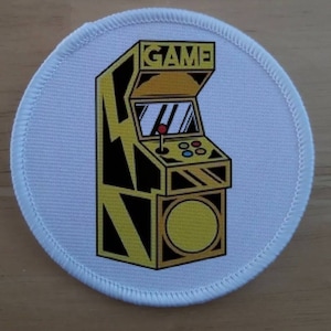 以下が含まれることがあります： 黒と黄色のアーケードゲームのイラストが描かれた白い丸いパッチ。ゲームの上には「GAME」という文字があり、ジョイスティックとボタンがあります。