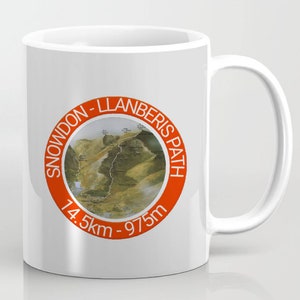 May include: White ceramic mug with an orange circle graphic featuring a map of the Snowdon-Llanberis Path. The text "Snowdon-Llanberis Path" is written around the circle, and the text "14.5km - 975m" is written below the circle.