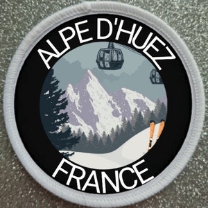 Op de afbeelding: Een zwarte en witte ronde patch met de tekst "ALPE D'HUEZ FRANCE" in wit. De patch toont een kleurrijke illustratie van een besneeuwd berglandschap met een skilift en skiërs.