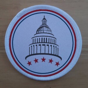 Op de afbeelding: Een ronde witte patch met een rood-wit-blauwe rand. In het midden van de patch bevindt zich een zwart-witte afbeelding van het Amerikaanse Capitool met vijf rode sterren eronder.