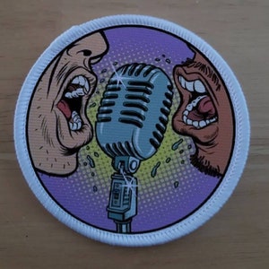 Può includere: Una toppa bianca con uno sfondo viola che presenta due volti di cartoni animati che cantano in un microfono vintage. Il microfono è argentato con una base nera e uno sfondo giallo e bianco.