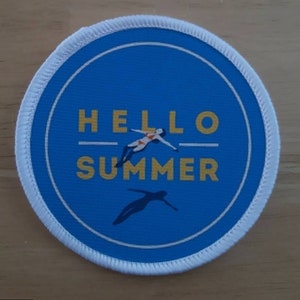Pode incluir: Um remendo redondo azul e branco com o texto "HELLO SUMMER" em letras amarelas. O remendo apresenta a silhueta de uma pessoa mergulhando na água.