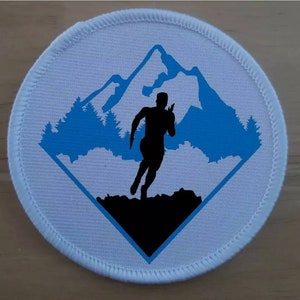 Puede incluir: Un parche bordado circular con fondo blanco y un borde en forma de diamante azul. Dentro del diamante hay una silueta azul de una cadena montañosa y pinos, con la silueta negra de una persona corriendo en el primer plano.